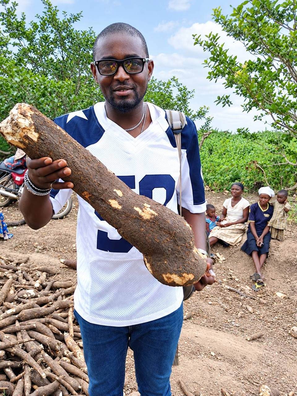 Holding a freshly harvested cassava root, with farmers gathered behind